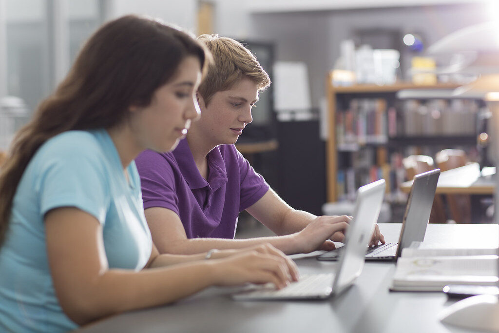 Dois estudantes sentados lado a lado em uma mesa de biblioteca, cada um trabalhando em seu próprio laptop.