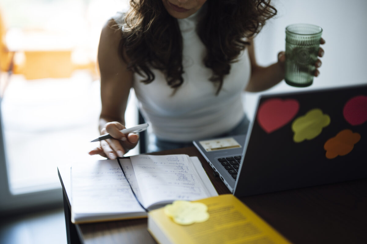Una mujer escribe en un cuaderno físico junto a un portátil abierto sobre un escritorio, con un libro y una nota adhesiva cerca, el tipo de notas mixtas en papel y digital que una herramienta de espacio de trabajo como Notion está pensada para organizar
