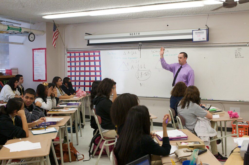 A teacher writing on a whiteboard at the front of a US high school classroom while students work at their desks.